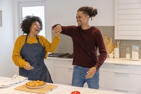 Playful couple laughing and dancing in bright modern kitchen while baking homemade pie