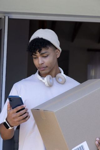 African american man holding package checking smartphone at doorway