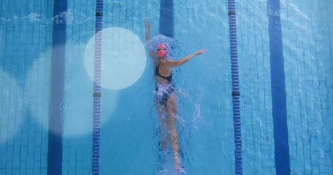 Backstroke athlete cutting through sunlit outdoor pool wearing pink cap, goggles, black swimsuit