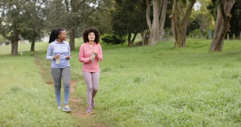 Two diverse women walking on park trail holding reusable cups and chatting in activewear