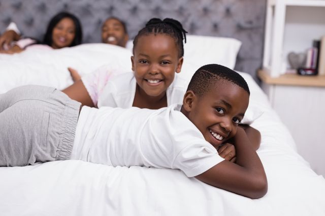 This image shows two smiling siblings lying on a bed with their parents in the background, creating a warm and joyful family atmosphere. Ideal for use in family-oriented advertisements, parenting blogs, lifestyle articles, and promotional materials for home and family products.