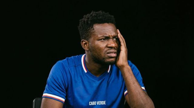 Pressing face and wincing, man wearing cabo verde soccer jersey against black backdrop