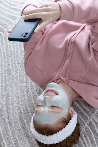 Young woman relaxing with facial mask and scrolling smartphone while lying on carpet