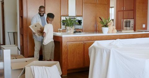 African American father and son packing moving boxes in modern kitchen with teddy bear