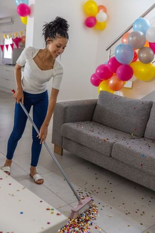 Teenage girl sweeping confetti after party in bright living room with balloons and broom