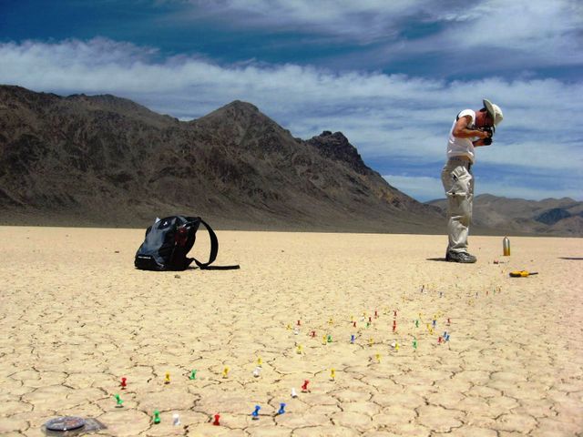 Image shows a scientist photographing a trail marked by pushpins in a dry lake bed at Racetrack Playa. The scientist wears a hat and a white outfit, with a black backpack on the ground. Suitable for illustrating scientific research, geological studies, desert landscapes, or NASA projects. Ideal for content related to environmental science, landscape photography, or educational materials about natural phenomena.