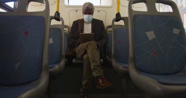 Mature man sitting alone on a public bus wearing a surgical mask and reading a tablet. Could be used for articles or advertising related to public transportation, health and safety measures, technology use among seniors, commuting, or urban living.