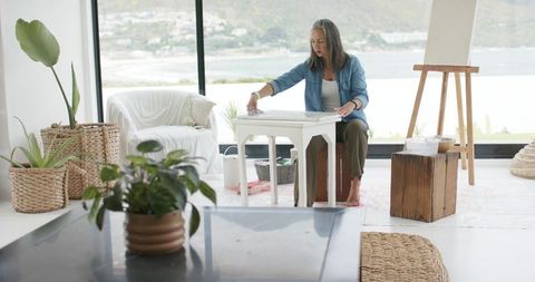 Senior woman painting small side table in sunlit modern living studio, diy home decor