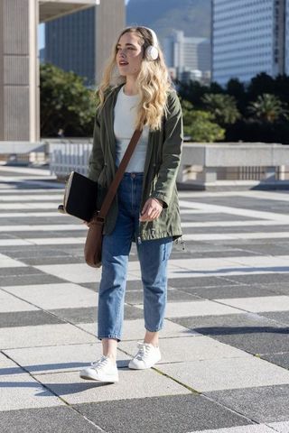 Young woman walking with skateboard and headphones across geometric plaza wearing green parka