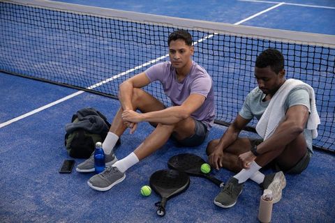 Male athletes resting on blue pickleball court after training, paddles, balls, hydration