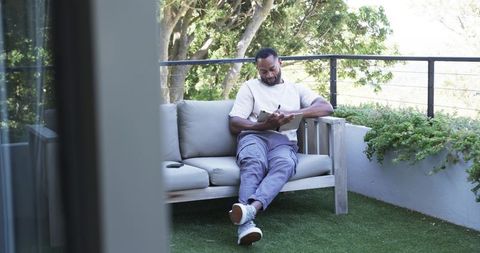 Mature man writing journal on balcony sofa, relaxing with smartphone and greenery