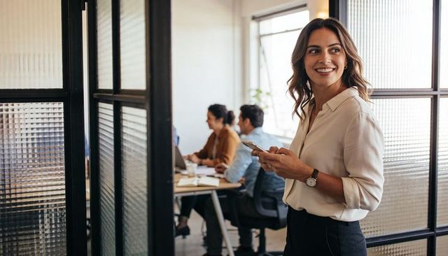 Smiling businesswoman holding smartphone outside glass office with team collaborating inside