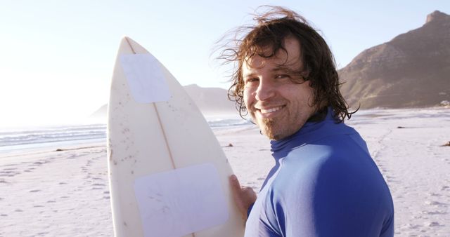 A Caucasian man in his young adulthood smiles while holding a surfboard on a sunny beach, with copy space. His wetsuit and surfboard suggest he's enjoying a day of water sports.