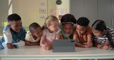 Multiracial children leaning over tablet collaborating and learning together in classroom