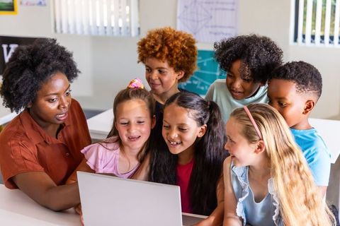 Diverse elementary students and teacher collaborating around laptop in bright classroom
