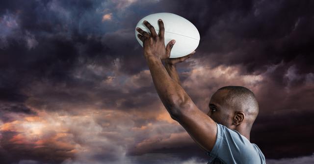 This shot captures a man holding a rugby ball against a dramatic cloudy sky. It is perfect for use in advertisements, blog posts about sports or motivation, or as part of a larger collection focusing on athletes or outdoor activities.