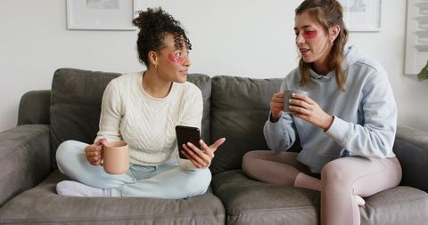 Friends enjoying cozy self-care night on couch with eye gel patches and hot drinks