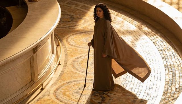 Standing woman holding cane wearing flowing robe with cape in sunlit marble atrium with mosaic floor