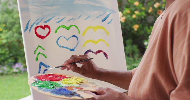 The artist is creating a vibrant, dynamic artwork filled with colorful hearts and birds on an easel using a paintbrush and palette. The setting is outdoors, with green and floral backdrop, suggesting a peaceful and inspiring environment for artistic endeavors. This can be used in websites or promos focused on art, creativity, hobbies, and leisure activities in nature.