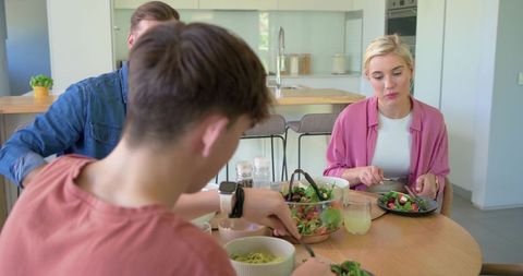 Young family sharing healthy salad dinner around round wooden table in modern kitchen