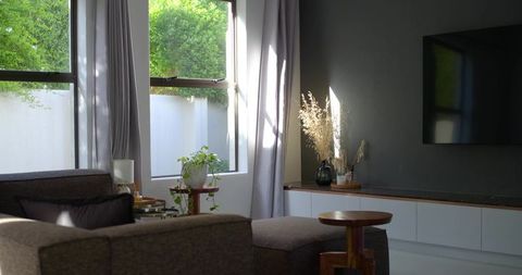 Sunlit minimalist living room with gray sectional, ottoman, tall windows and greenery
