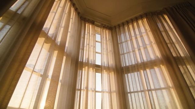 Filtering sheer curtains through bay window, casting muntin shadows and warm sunlight glow
