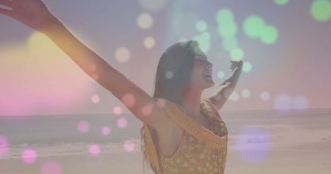 A woman wearing a yellow floral dress stands on the beach, spreading her arms wide. The scene features delightful colored bokeh lights, evoking a sense of freedom and optimism. Ideal for use in lifestyle blogs, summer travel showcases, relaxation themes, and products focused on well-being and happiness.