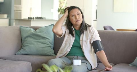 Mature woman monitoring blood pressure at home on sofa using portable cuff and monitor