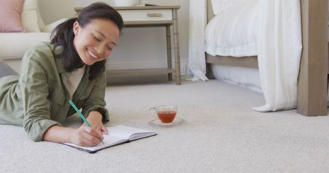 The image shows an Asian woman lying comfortably on the carpet, smiling while writing in a notebook, representing creativity and relaxation at home. Ideal for use in lifestyle content, articles about leisure activities, creative thinking, home relaxation inspiration, or promotional materials for writing retreats or mental well-being campaigns.
