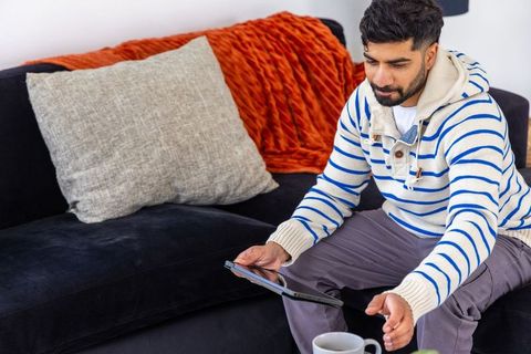 Man sitting on sofa holding tablet and reaching for coffee mug wearing striped knit hoodie
