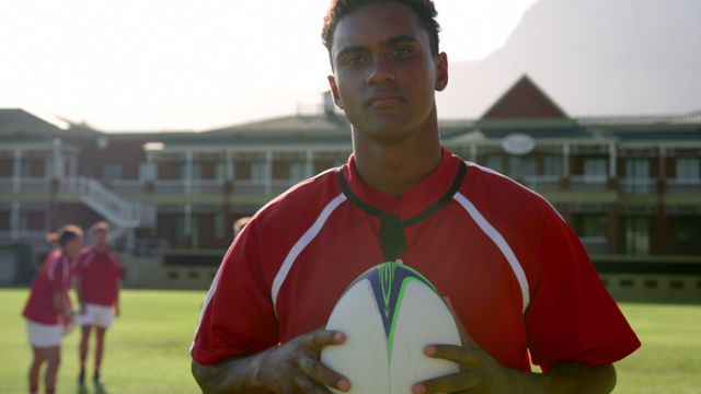 Confident male rugby player standing and holding a rugby ball on a sunny field with teammates in background. Ideal for branding athletic programs, sports event promotions, motivational content, fitness training materials, and sports equipment advertisements.