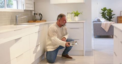 Senior man kneeling and holding stainless pet bowl in modern bright white kitchen at home