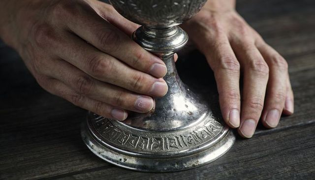 Cradling hands holding ornate silver chalice base on rustic wooden tabletop closeup
