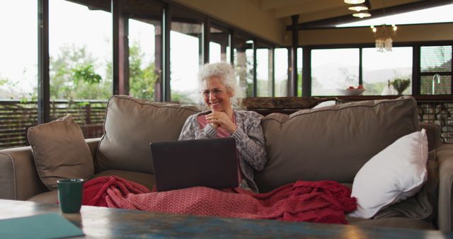 Senior woman is relaxing on a couch at home, using a laptop. She is draped with a red blanket and is smiling, showing contentment. The setting features large windows that provide ample natural light and a spacious, modern living room. Ideal for depictions of home life, elderly care, online communication, remote work, and comfortable living.