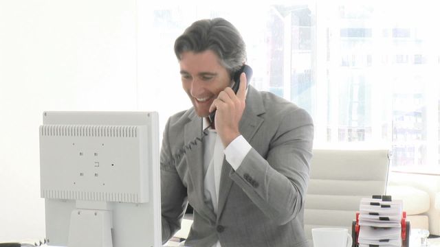 Businessman talking on the phone while using a computer at modern office desk, smiling. Perfect for content on corporate culture, teamwork, executive roles, business consulting, and workplace efficiency.