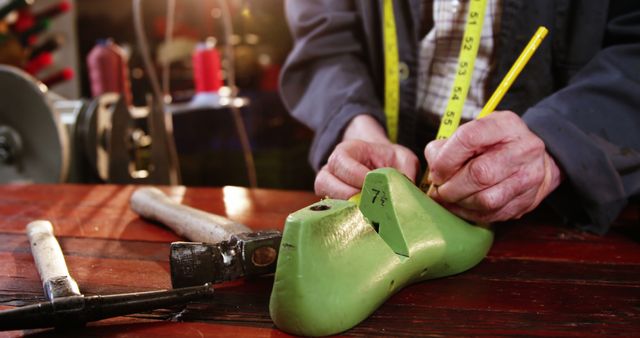 Image features a cobbler's hands crafting shoes in a workshop. Cobbler is using a measuring tape on a green shoe mold. The scene captures traditional craftsmanship with tools scattered on a wooden table. This visual can be used for articles about traditional trades, craftsmanship, handmade products, and artisanal skills. It is suitable for use in marketing campaigns, blogs, magazines, and educational content related to shoemaking and artisan crafts.