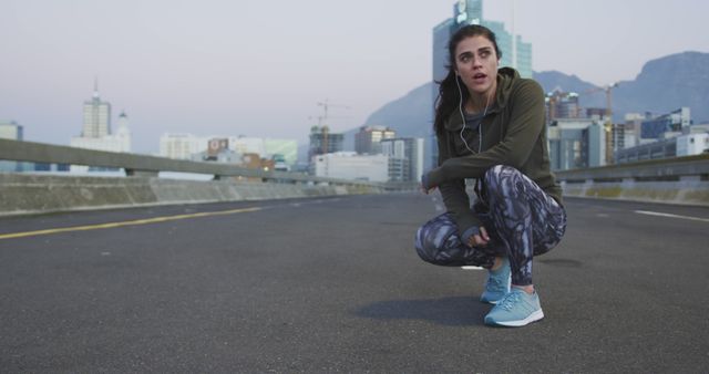 Caucasian woman wearing earphones, kneeling and taking break looking side with copy space in morning. Excercising, jogging, music, sport and healthy lifestyle, unaltered.