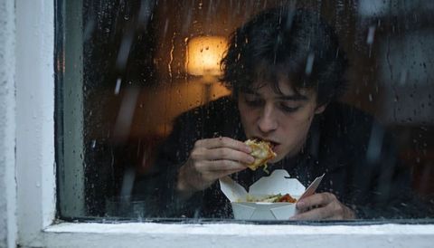 Young man eating takeout behind rainy window during late-night storm warm lamp glow
