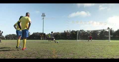 Player taking penalty shot with goalkeeper diving on sunlit grass pitch and teammates watching