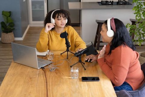 Diverse women recording cozy home podcast at kitchen table with laptop and microphones