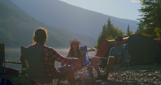 Friends enjoying scenery and conversation at lakeside campsite with mountain backdrop. Ideal for promoting camping equipment, outdoor adventure advertisements, travel blogs, lifestyle magazines, and serene nature settings.