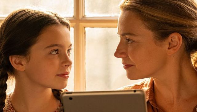 Mother and daughter sharing warm backlit moment by window with tablet, intimate portrait