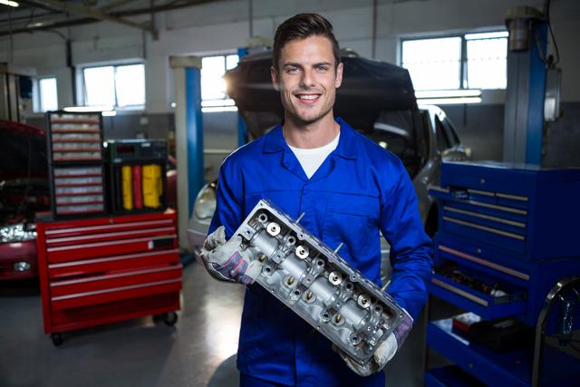 Mechanic in blue uniform smiling while holding a car engine part in a well-equipped repair garage. Ideal for use in automotive service advertisements, mechanic training materials, and promotional content for car repair shops.