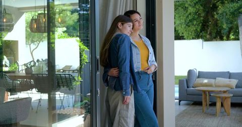 Diverse female couple embracing in sunlit modern home doorway with patio view