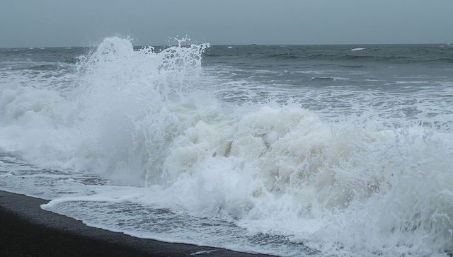 Waves crashing on dark sand shoreline sending tall vertical spray under stormy sky