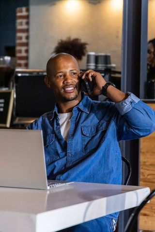 African american man working remotely in cafe on laptop while talking on smartphone
