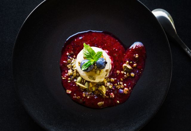 Elegant presentation of a gourmet dessert featuring vanilla ice cream on a berry sauce, topped with crunchy nuts and a mint leaf on a black plate. Ideal for use in food blogs, restaurant menus, culinary magazines, and social media posts celebrating fine dining and gourmet cuisine.