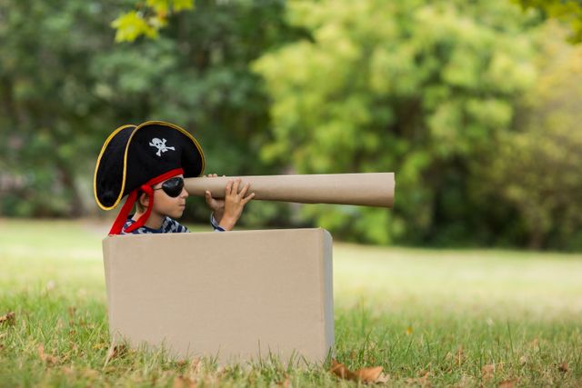 Young boy dressed as a pirate, sitting in a cardboard box, holding a telescope, enjoying imaginative play in a park. Ideal for use in articles or advertisements about childhood, creativity, outdoor activities, and imaginative play.
