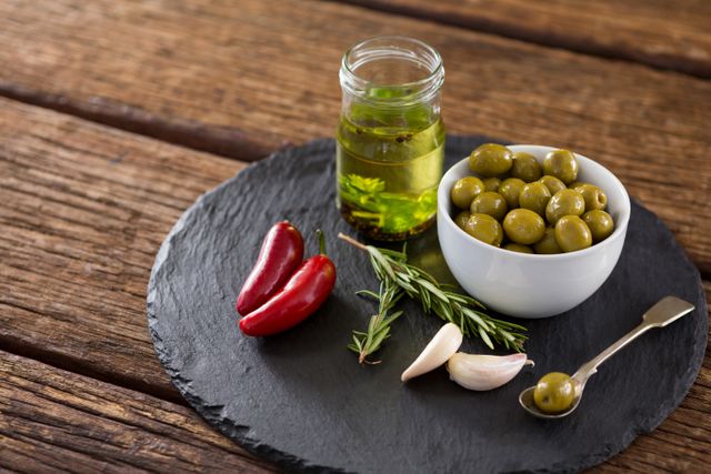 Green olives in white bowl with jar of olive oil, red chilies, garlic, and rosemary on rustic wooden table. Ideal for illustrating Mediterranean cuisine, healthy eating, and gourmet food preparation. Perfect for food blogs, recipe websites, and culinary magazines.