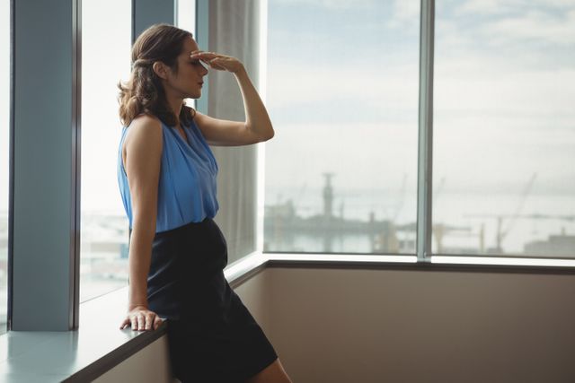 Businesswoman standing near window in office, looking stressed and thoughtful. Ideal for illustrating workplace stress, corporate environments, and professional challenges. Useful for articles on business, mental health, and career development.